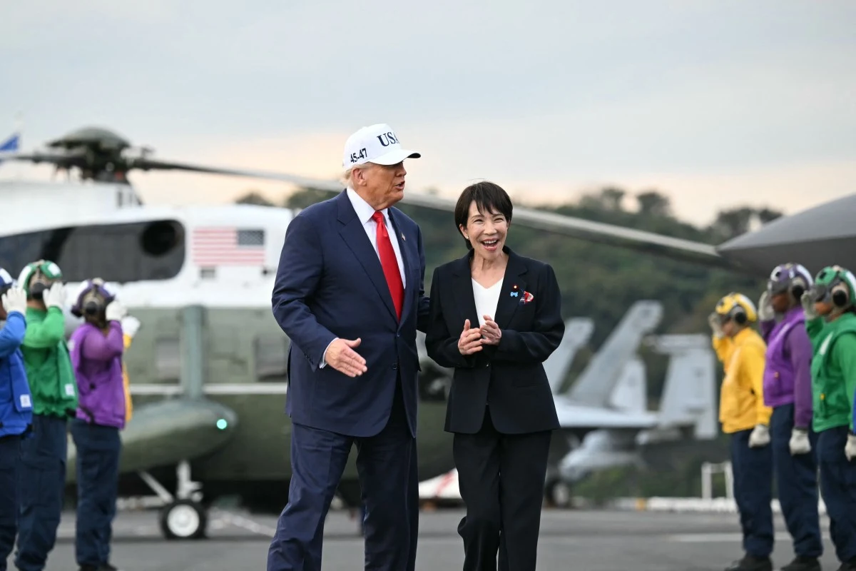 Prezydent USA Donald Trump i premier Japonii Sanae Takaichi Dwóch elegancko ubranych ludzi spaceruje wspólnie i rozmawia na tle śmigłowca z amerykańską flagą, wokół nich stoją osoby w kolorowych uniformach, przypominających obsługę lotniska bądź pokładu lotniskowca.