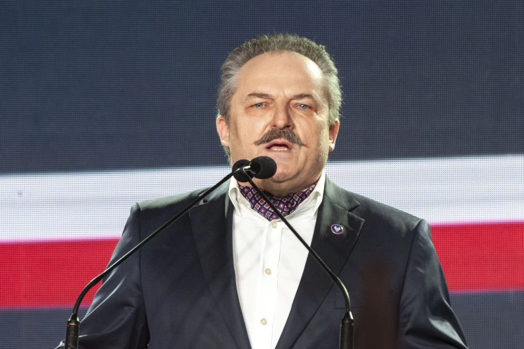 Mężczyzna w garniturze oraz apaszce przemawia do mikrofonu na tle dużej flagi w barwach biało-czerwonych.