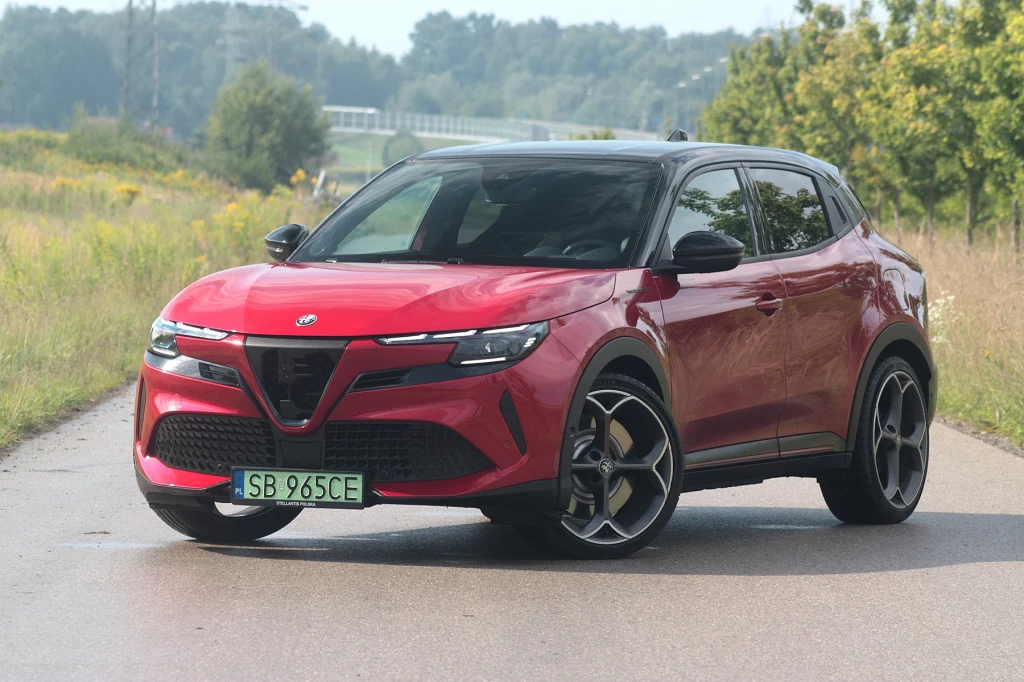 Czerwony samochód typu SUV z czarnym dachem oraz sportowymi felgami stoi na asfaltowej drodze otoczonej zielenią, w tle widoczne drzewa i lekko zamglony krajobraz.