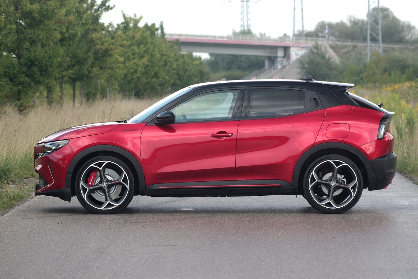 Bez punktu odniesienia Junior wygląda na znacznie większy niż w rzeczywistości Czerwony samochód typu SUV zaparkowany na asfalcie w naturalnym otoczeniu, na tle zieleni i mostu w oddali.