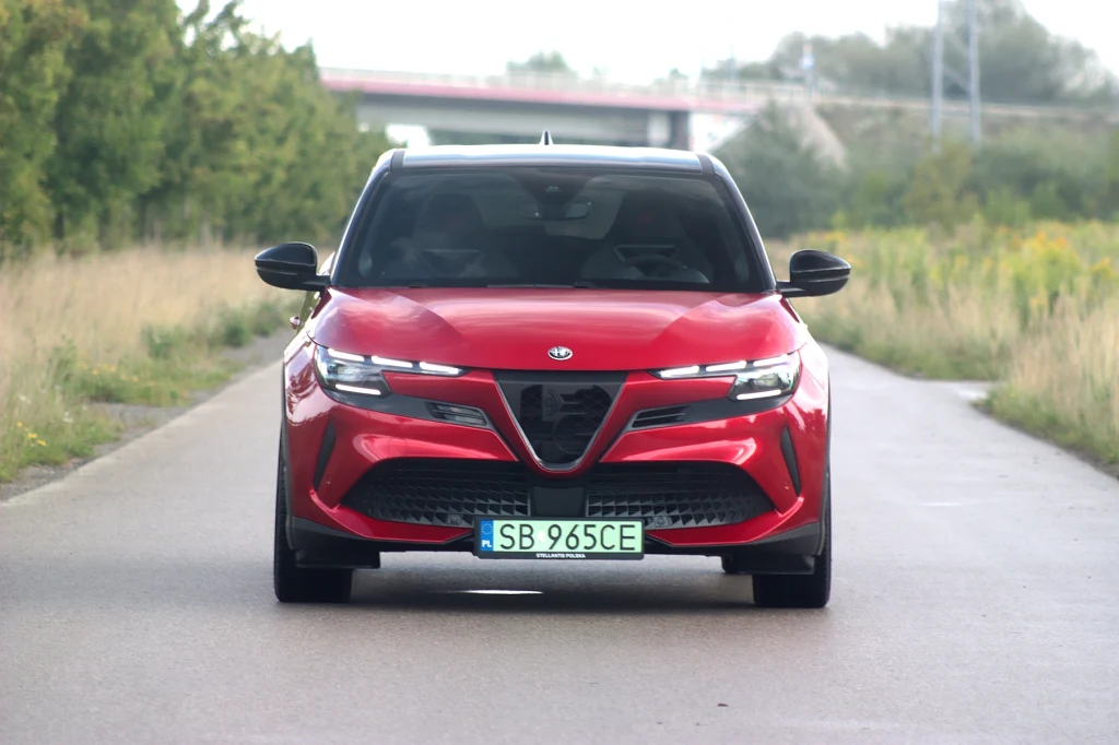 Czerwony samochód Alfa Romeo Tonale widoczny z przodu, stojący na pustej, asfaltowej drodze otoczonej zielenią i niską roślinnością, w tle wiadukt.