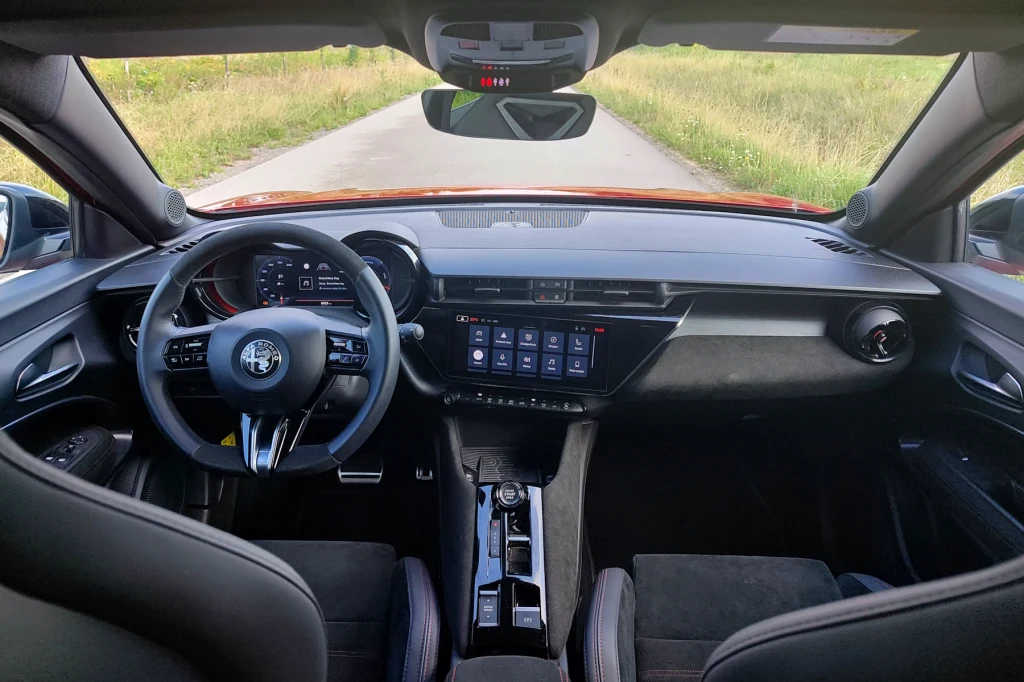 Widok wnętrza nowoczesnego samochodu osobowego marki Alfa Romeo z perspektywy tylnego siedzenia, centralnie umieszczona kierownica, cyfrowy kokpit, duży ekran dotykowy na konsoli środkowej, obecność dwóch przednich foteli, naturalne światło wpadające p...