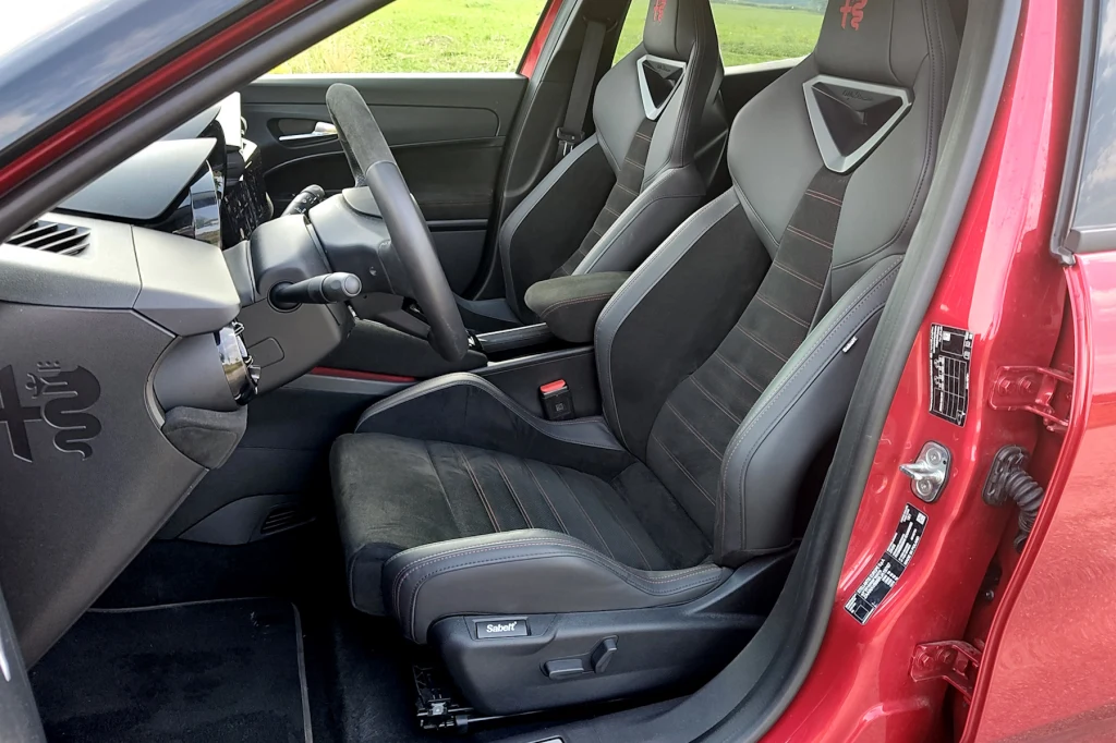 Kokpit sportowego samochodu z charakterystycznymi kubełkowymi fotelami z logo, czarną tapicerką i czerwonymi przeszyciami, widoczne szczegóły wykończenia wnętrza, otwarte drzwi oraz fragment deski rozdzielczej i kierownicy.