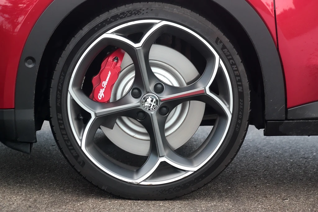 Duże aluminiowe felgi z charakterystycznym logo Alfa Romeo na środku, czerwony zacisk hamulcowy z napisem marki oraz opona na tle szarego asfaltu. Część czerwonej karoserii widoczna w tle.