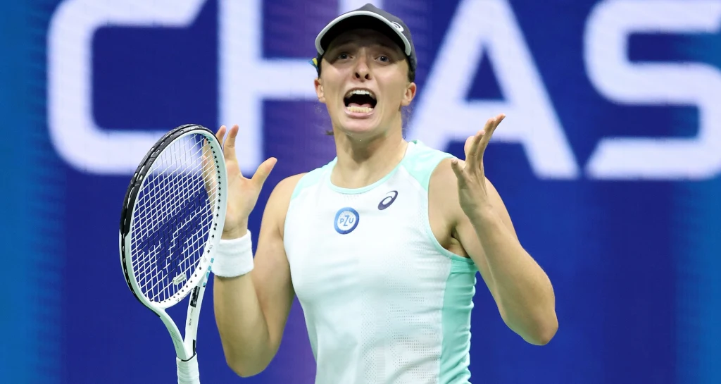 Tenisistka w sportowym stroju z rakietą w ręku podczas meczu na korcie, wyrażająca silne emocje poprzez gestykulację i mimikę twarzy, w tle nieostre logo sponsora.