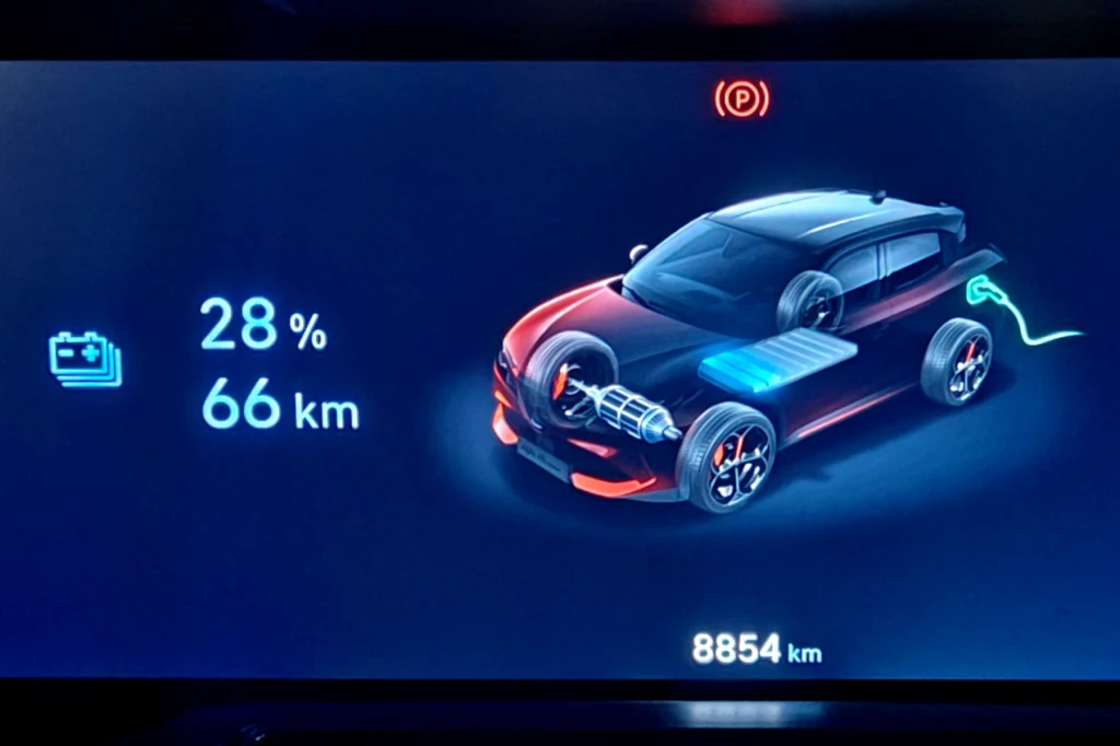 Wyświetlacz samochodu elektrycznego pokazuje graficzną wizualizację pojazdu z zaznaczonymi elementami systemu elektrycznego, ikoną wtyczki ładującej, poziomem naładowania akumulatora wynoszącym 28% oraz szacowanym zasięgiem 66 km i przebiegiem całkowit...