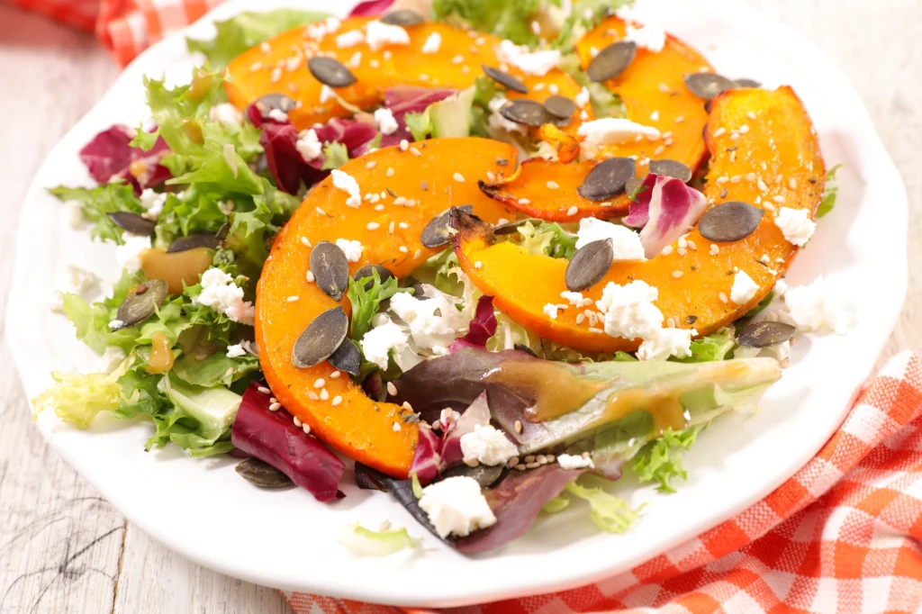 Przepis na sałatkę z dyni można dowolnie modyfikować Kolorowa sałatka z mieszanką świeżych liści sałat, podprażonymi pestkami dyni, kawałkami sera feta oraz prażonymi plasterkami dyni, ułożona na białym talerzu i ozdobiona sezamem.