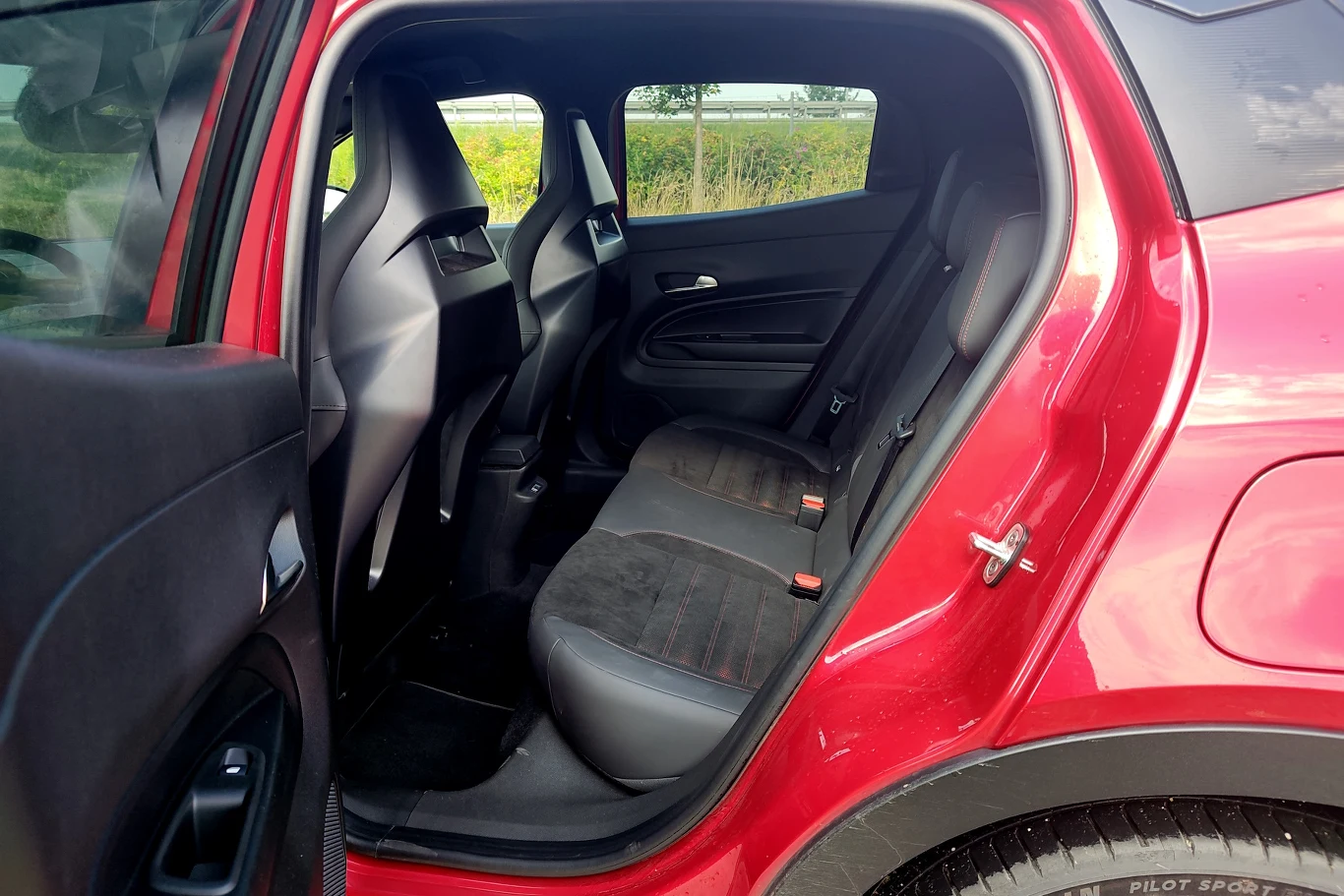 Alfa Romeo Junior Otwarta tylna para drzwi czerwonego samochodu ukazująca czyste i zadbane tylne siedzenia, wykończone ciemną tapicerką z czerwonymi przeszyciami. W tle widoczne zielone otoczenie.