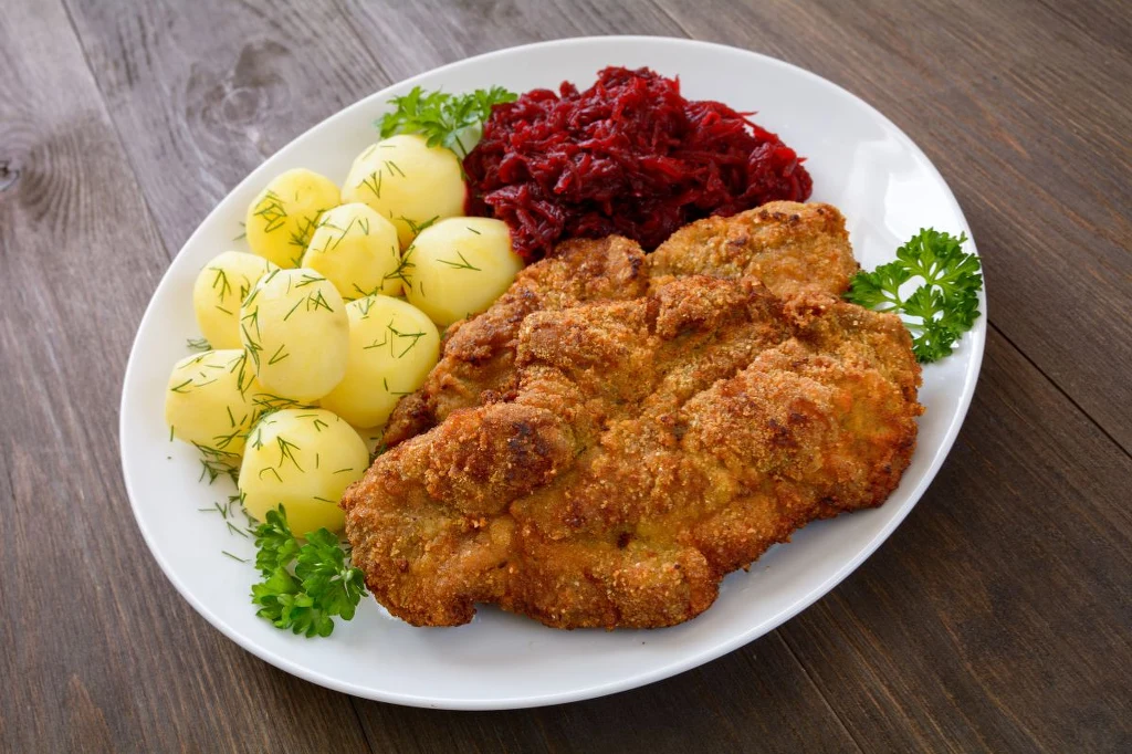 Ulubiony obiad milionów Polaków nie jest łaskawy dla jelit Chrupiący kotlet schabowy w panierce, porcje młodych ziemniaków z koperkiem oraz buraczki na ciepło, podane na białym talerzu, ozdobione gałązkami natki pietruszki, ustawione na drewnianym stole.