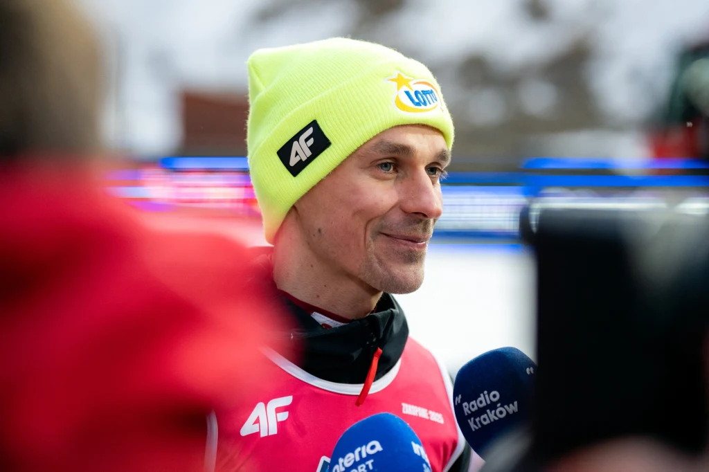 Piotr Żyła Sportowiec w żółtej czapce i czerwonym stroju udziela wywiadu mediom, otoczony mikrofonami z logotypami stacji radiowych, w tle rozmyte zimowe otoczenie.