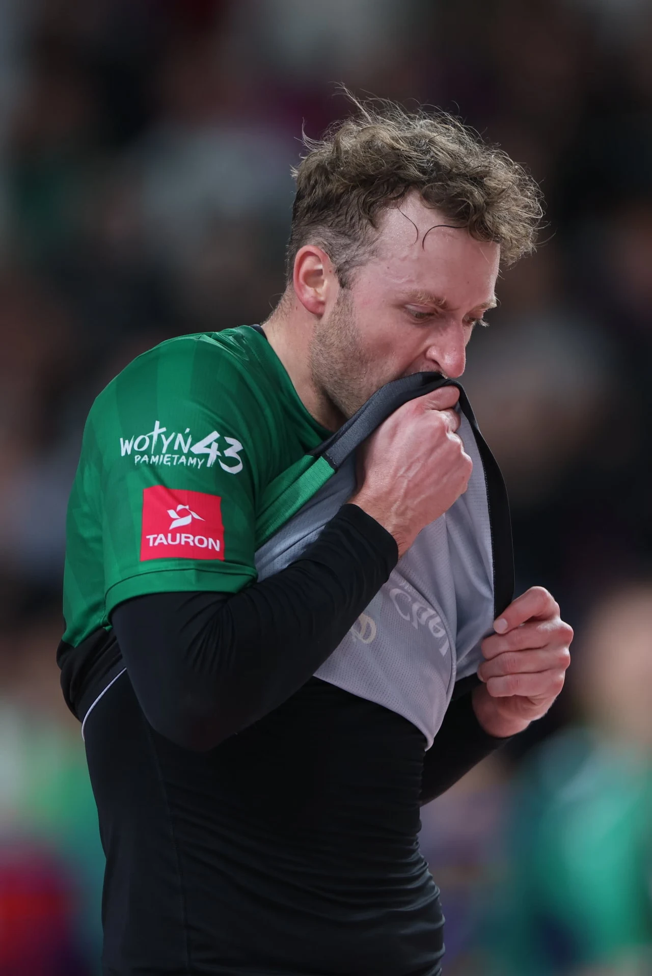 Siatkarz CHKS Chełm Sportowiec w zielonej koszulce trzyma i przyciska do twarzy swoją koszulkę, wyrażając silne emocje. Na ramieniu widoczny czerwony logotyp sponsora, wokół rozmyte tło z tłumem ludzi.
