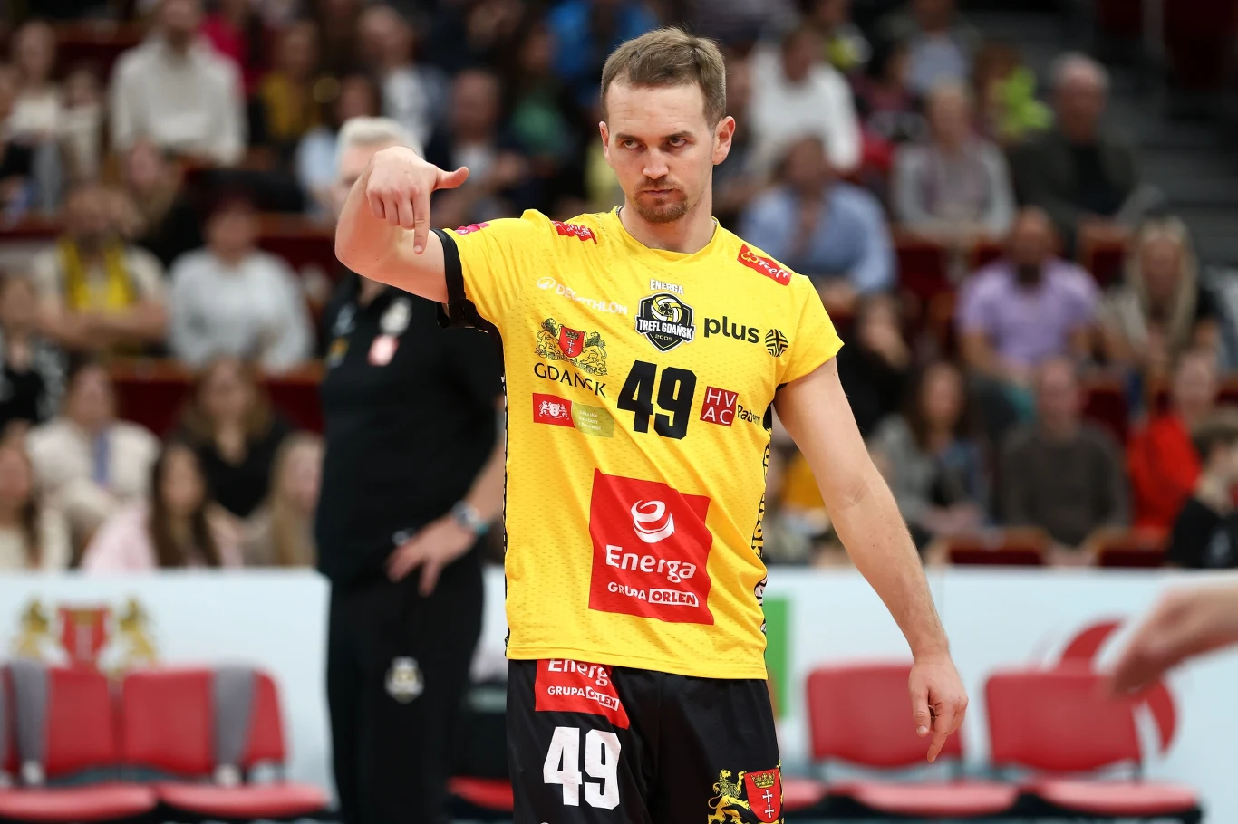 Joseph Worsley w barwach Energi Trefla Gdańsk Siatkarz w żółtej koszulce z numerem 49 na tle trybun podczas meczu, wykonujący gest ręką, w tle widoczni kibice siedzący na trybunach.