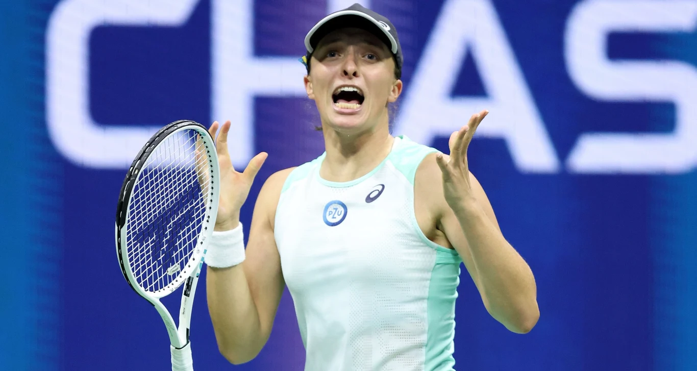 Iga Świątek Tenisistka w stroju sportowym z rakietą w ręku podczas meczu na korcie, z wyrazem emocji na twarzy, w tle rozmyte elementy hali z widocznym fragmentem napisu.