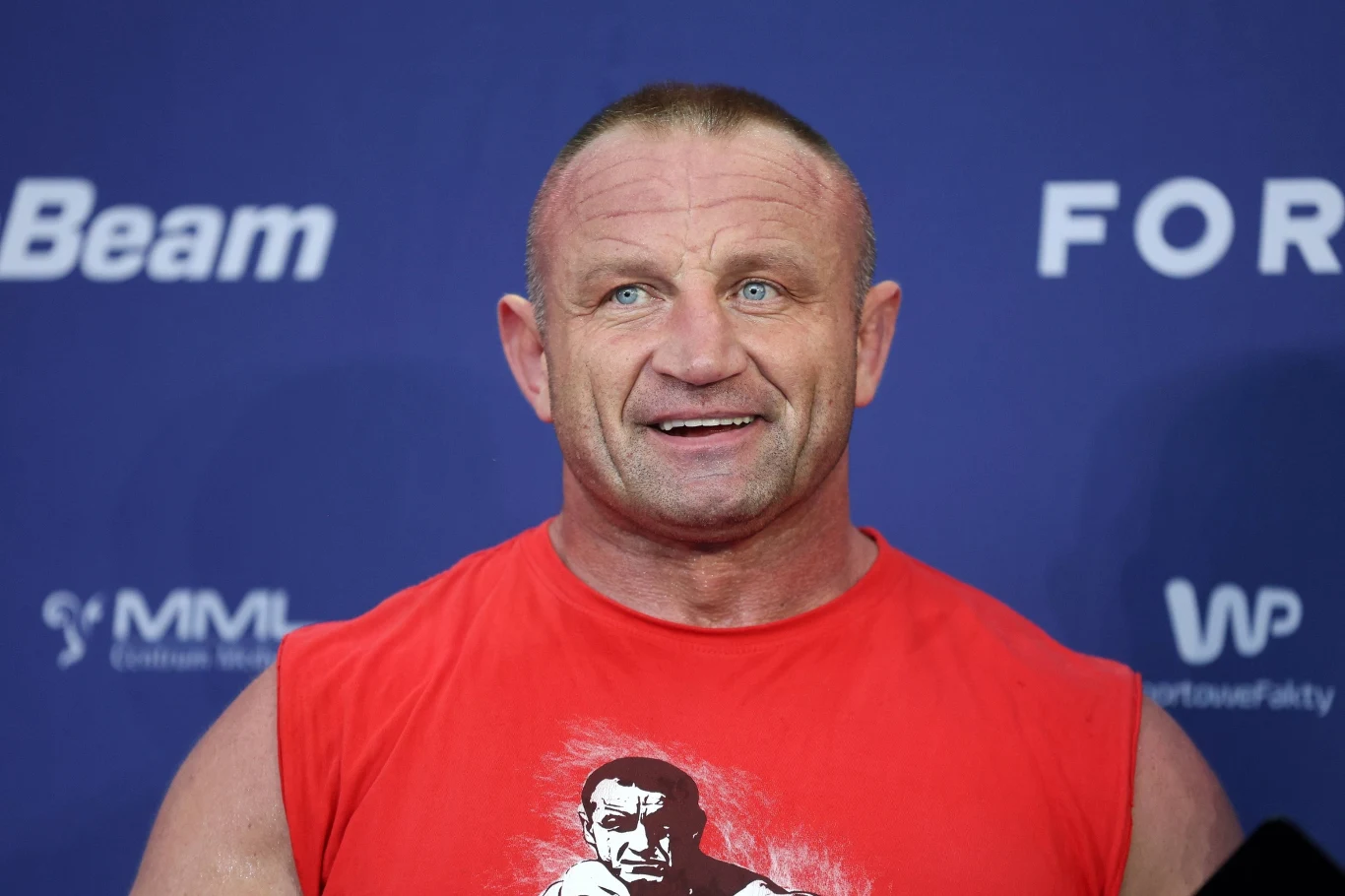 Mariusz Pudzianowski Mężczyzna o atletycznej sylwetce w czerwonej koszulce bez rękawów, z nadrukiem twarzy, stoi na tle niebieskiej ścianki z logotypami.