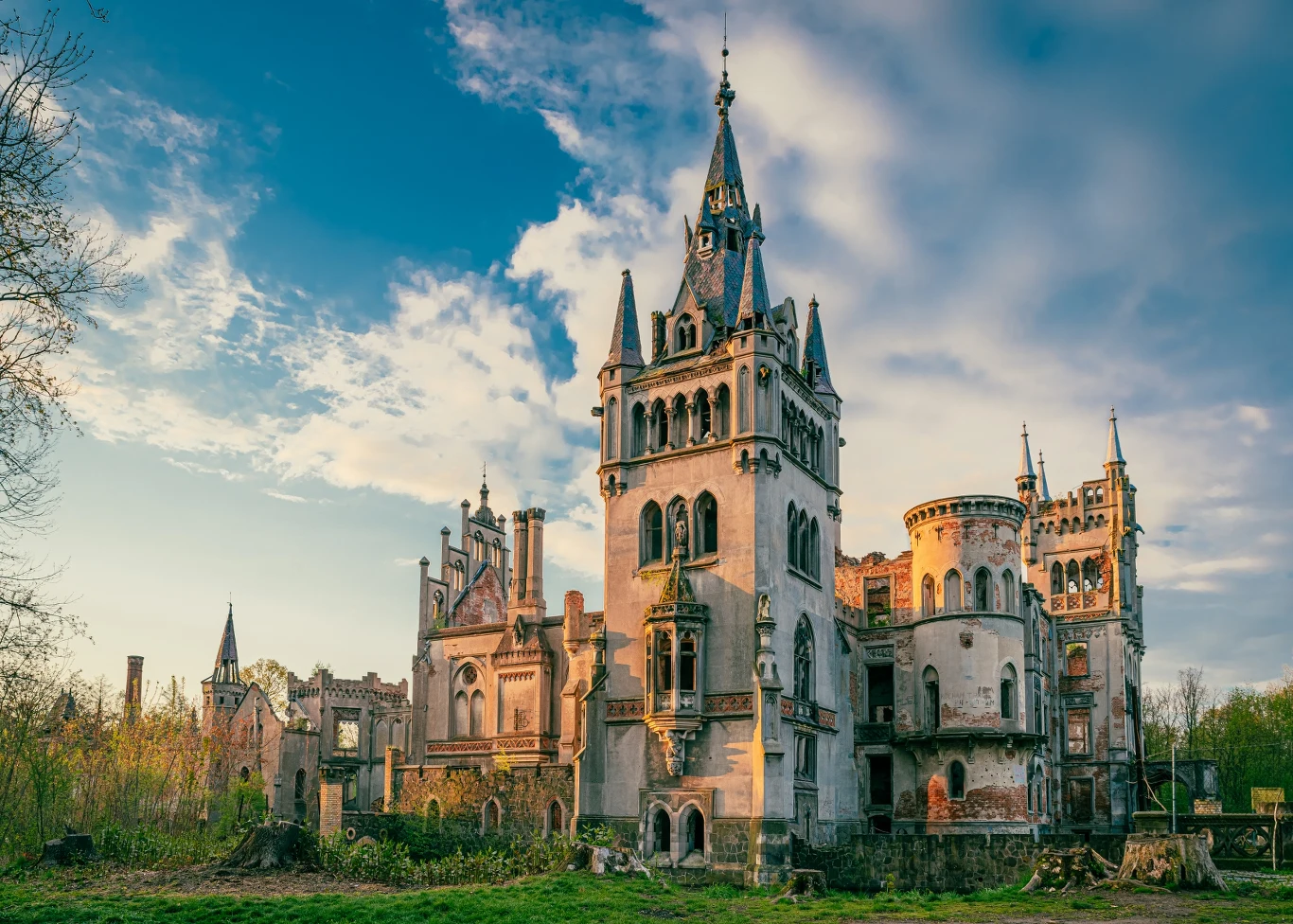 Zabytkowy zamek o wysokich, strzelistych wieżach i licznych oknach otoczony zarośniętym trawnikiem i drzewami, na tle niebieskiego nieba z chmurami.