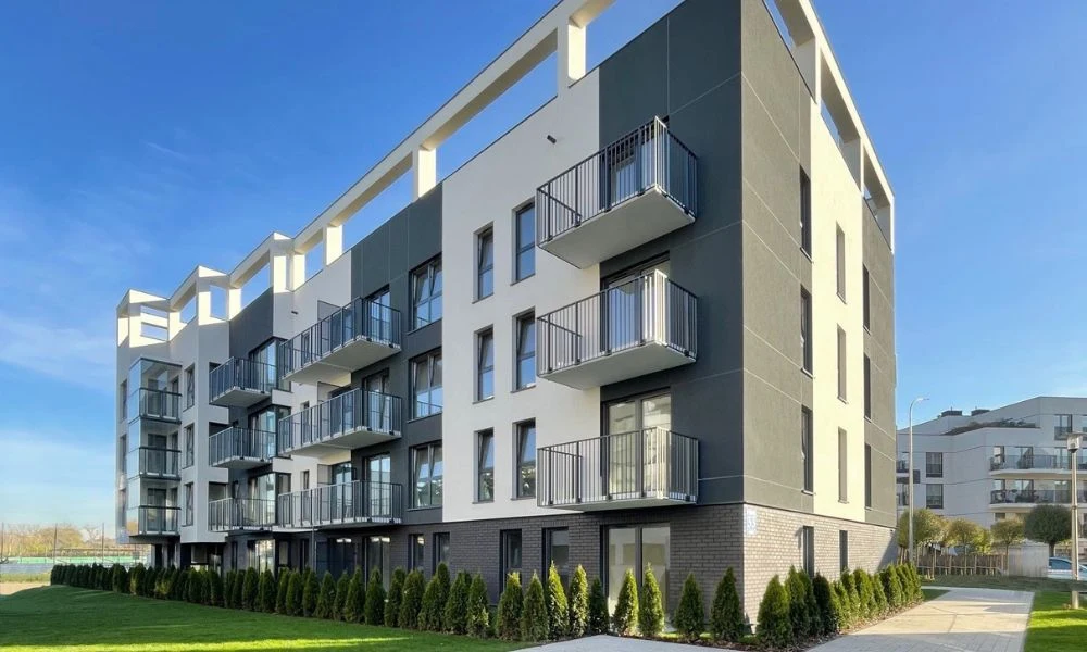 Deweloper sfinalizował osiedle w Starym Podgórzu w Krakowie Nowoczesny blok mieszkalny o jasnej elewacji z elementami grafitowymi, czterokondygnacyjny, z wieloma balkonami, otoczony zielenią i niskim żywopłotem, w tle widoczne inne zabudowania urbanistyczne.