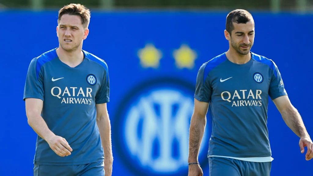 Dwóch piłkarzy w niebieskich koszulkach treningowych klubu Inter Mediolan spaceruje po boisku, w tle niebieska plansza z logo klubu i dwoma żółtymi gwiazdkami.