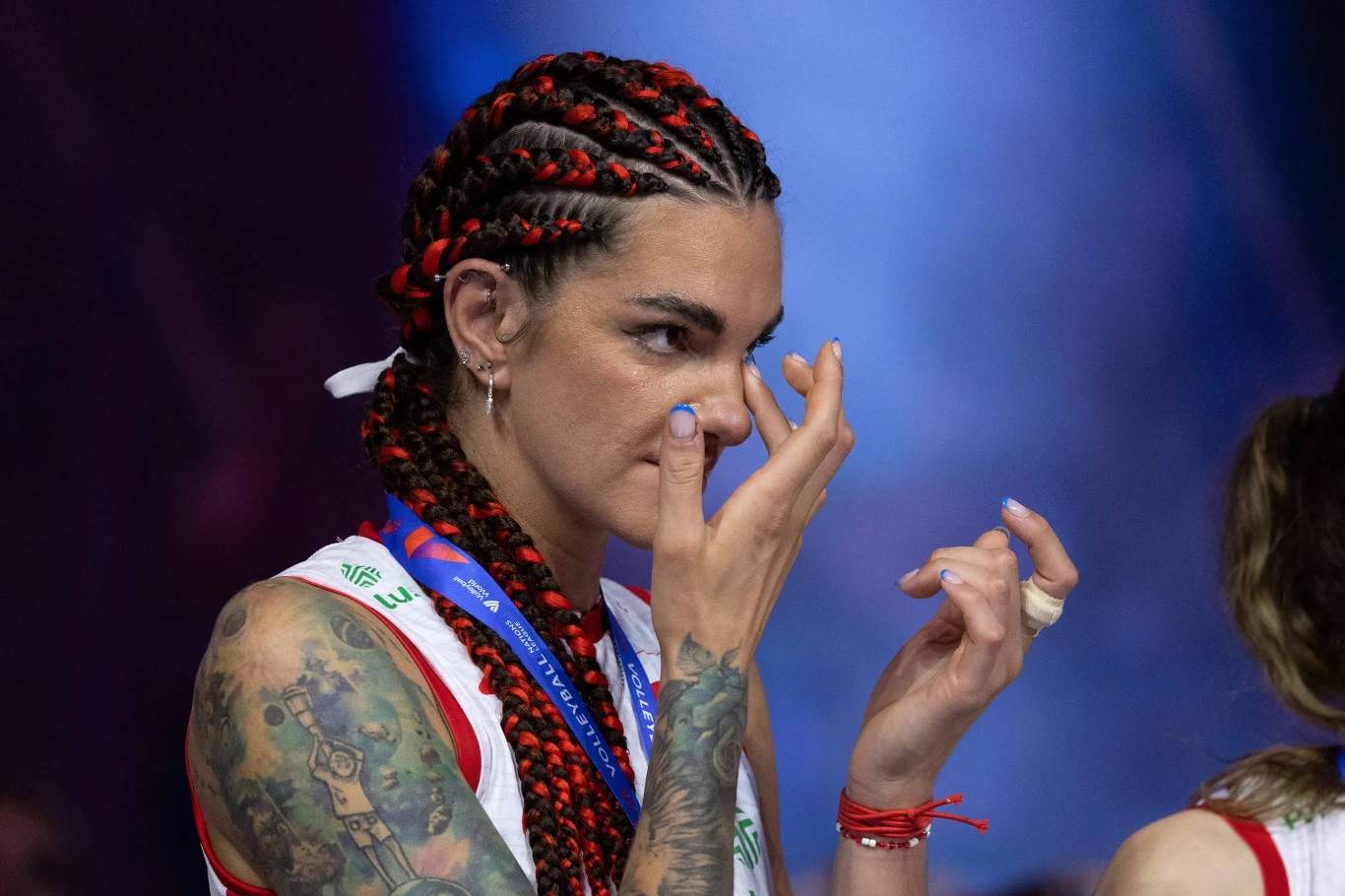 Malwina Smarzek Kobieta w sportowym stroju z medalem na szyi, z wyrazistymi tatuażami na ramieniu, poprawia makijaż lub ociera łzę, ma kolorowe warkoczyki oraz skoncentrowany wyraz twarzy, w tle widoczna niewyraźna postać.