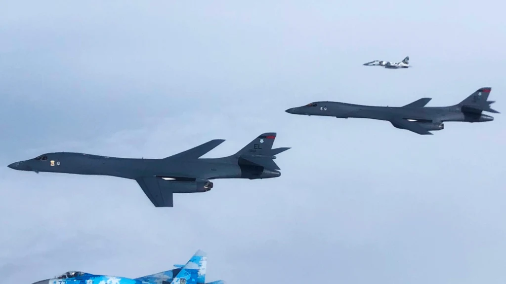 Para bombowców B-1 Lancer przeleciała tuż obok Caracas. To wymowny sygnał Trumpa dla Wenezueli (zdjęcie ilustracyjne) Dwa duże bombowce wojskowe lecą równolegle w powietrzu, w tle widoczny jest mniejszy myśliwiec oraz fragment samolotu o niebieskim ubarwieniu w lewym dolnym rogu.