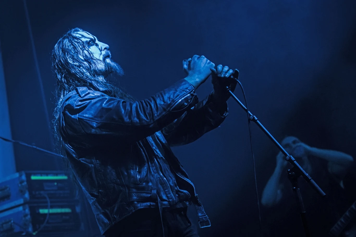 Gaahl stoi na czele grupy Gaahls Wyrd Wokalista zespołu metalowego trzyma mikrofon na scenie, ubrany w czarną skórzaną kurtkę, oświetlony niebieskim światłem scenycznym, z długimi, mokrymi włosami, w tle drugi muzyk i sprzęt muzyczny.