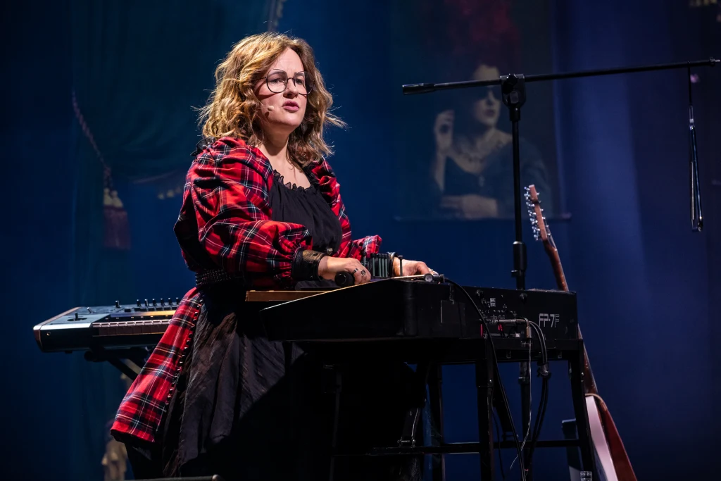 Kobieta w okularach siedzi przy klawiaturze elektronicznej na scenie, ubrana w czerwoną koszulę w kratę, z boku widoczna gitara oraz klawiatura, w tle ciemna dekoracja z obrazem kobiety.