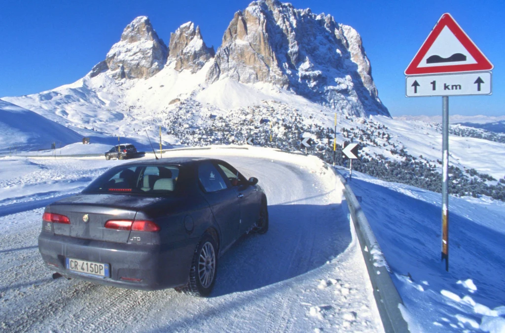 Samochód osobowy poruszający się zaśnieżoną krętą drogą w górach, obok znak ostrzegawczy o nierównościach na drodze, w tle ośnieżone szczyty i wyraźne skaliste formacje.