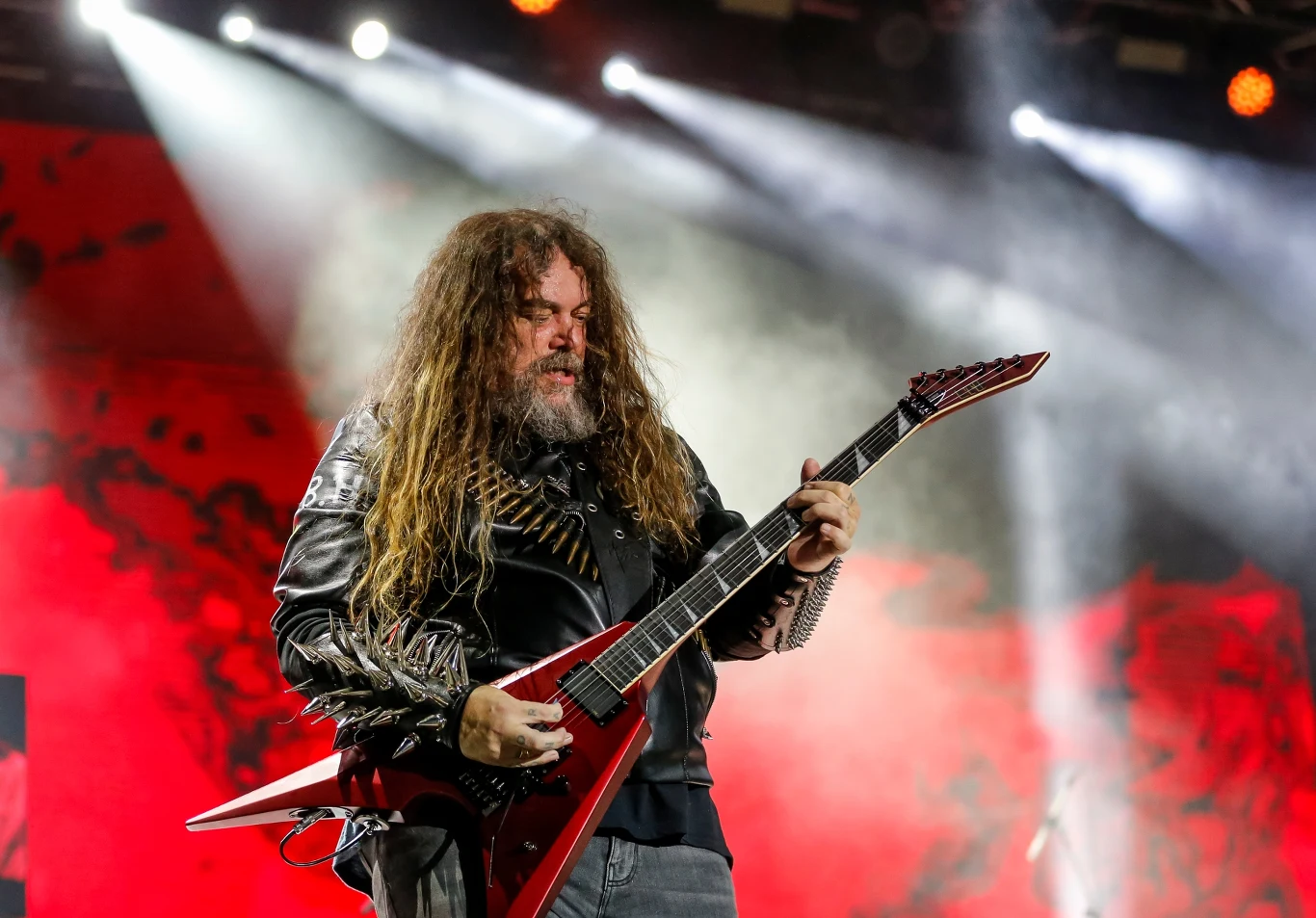 Max Cavalera stoi na czele grupy Soulfly Długowłosy muzyk w kurtce skórzanej z ćwiekami grający na czerwonej gitarze elektrycznej typu V na tle sceny oświetlonej reflektorami i czerwonym światłem.