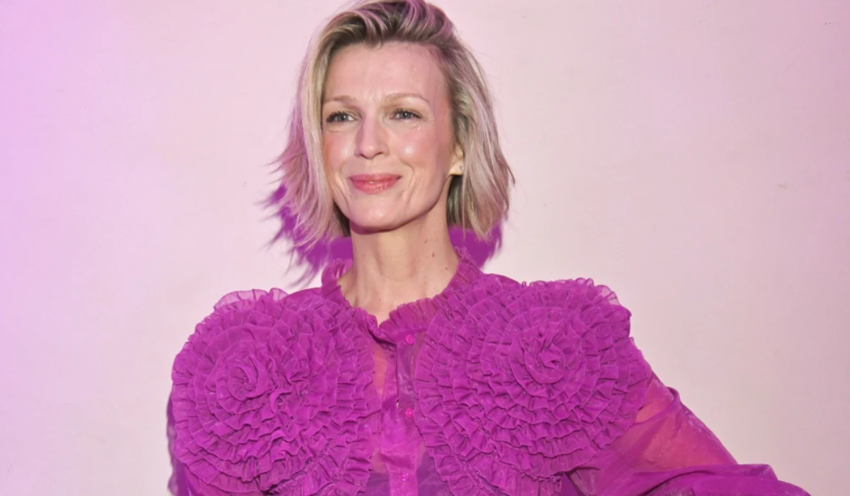 Magda Mołek Kobieta o jasnych, krótkich włosach w fioletowej, ozdobnej bluzce z marszczeniami i falbanami na tle jasnej ściany z delikatnym światłem fioletowych refleksów.