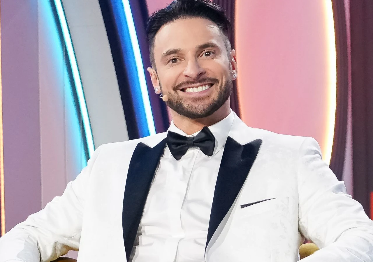 Piotr z "Warsaw Shore" w "Randce w ciemno" Uśmiechnięty mężczyzna w białym smokingu z czarnymi klapami oraz czarną muchą siedzi na żółtym fotelu na tle kolorowych, podświetlanych paneli.