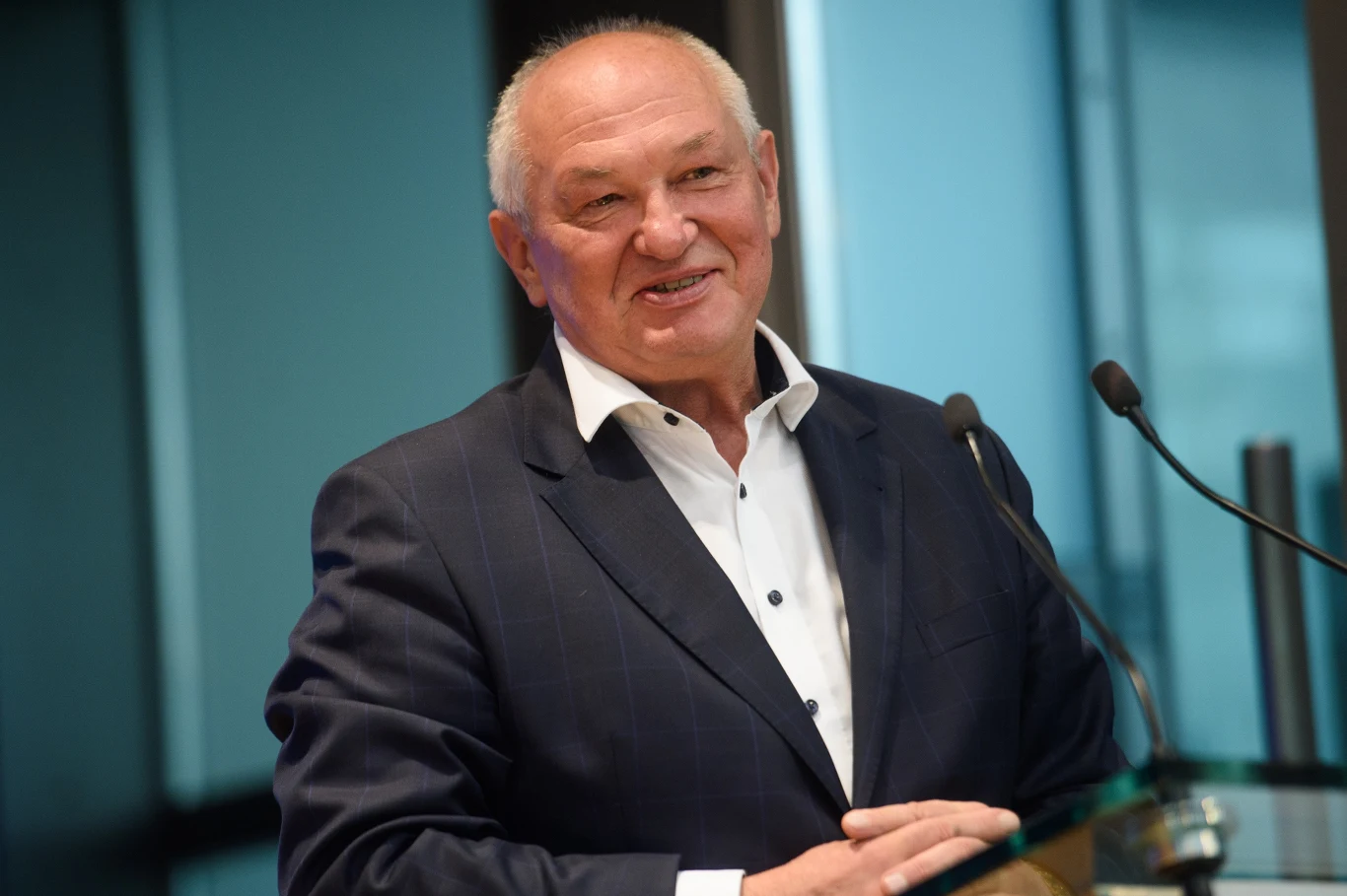 Waldemar Humięcki Starszy mężczyzna w garniturze stoi przy mównicy, uśmiecha się i mówi do mikrofonu. W tle widoczne są szklane ściany i nowoczesne wnętrze biurowe.