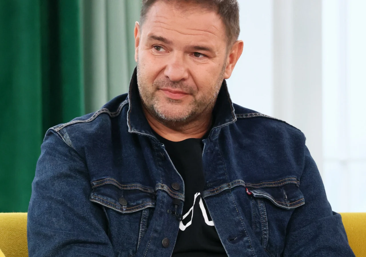 Tomasz Karolak Mężczyzna w średnim wieku siedzi na żółtej sofie, ubrany w ciemną dżinsową kurtkę i czarną koszulkę, z wyrazem łagodnej zamyślonej twarzy. W tle widoczne zielone zasłony.