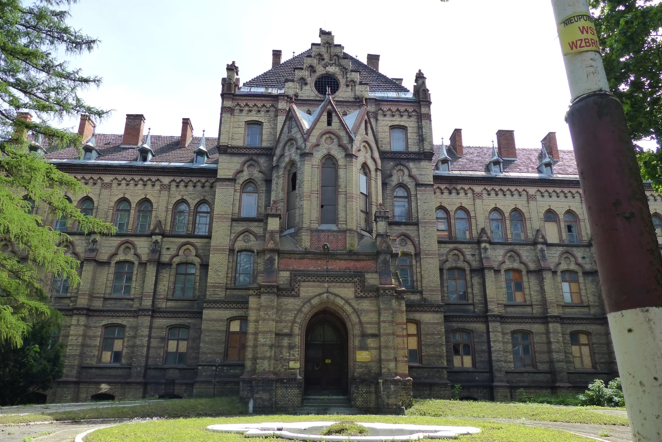 Zabytkowy, wielopiętrowy budynek z czerwonej cegły z licznymi detalami architektonicznymi i ozdobnymi oknami, otoczony zielenią drzew. Na pierwszym planie wejście łukowe i rozległy trawnik przed gmachem.