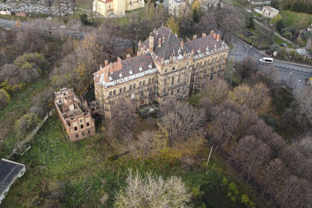 Pałac w Mokrzeszowie widziany z lotu ptaka Duży, zabytkowy pałac z czerwonej cegły otoczony zarośniętym parkiem, obok znajduje się zrujnowany budynek, w tle widoczne ulice oraz kilka domów jednorodzinnych.
