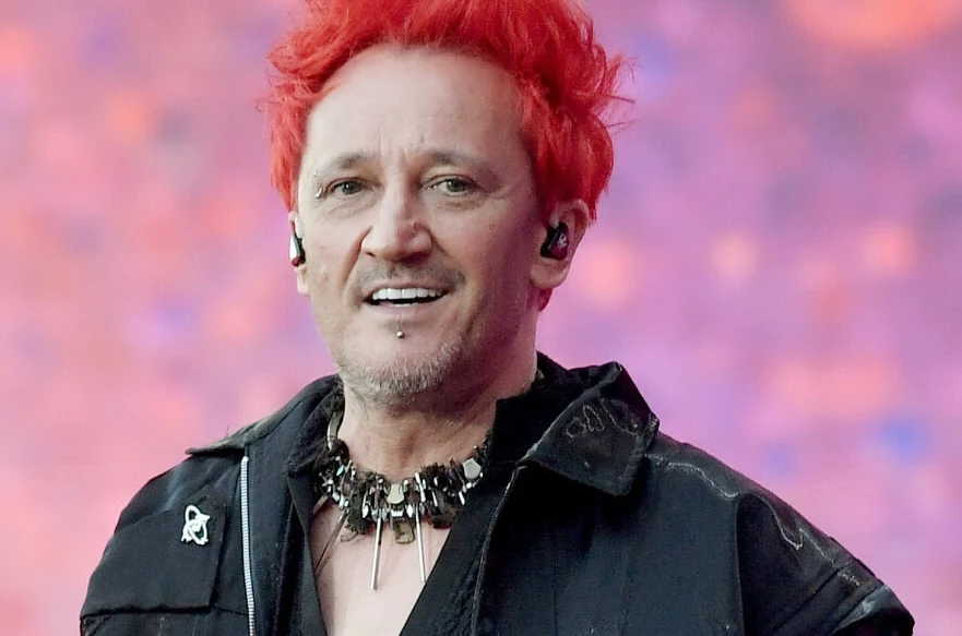 Michał Wiśniewski Mężczyzna z intensywnie czerwonymi włosami, uśmiechnięty, ubrany w czarną kurtkę z metalowym, ozdobnym naszyjnikiem i słuchawkami dousznymi, na tle rozmytego różowo-fioletowego tła.