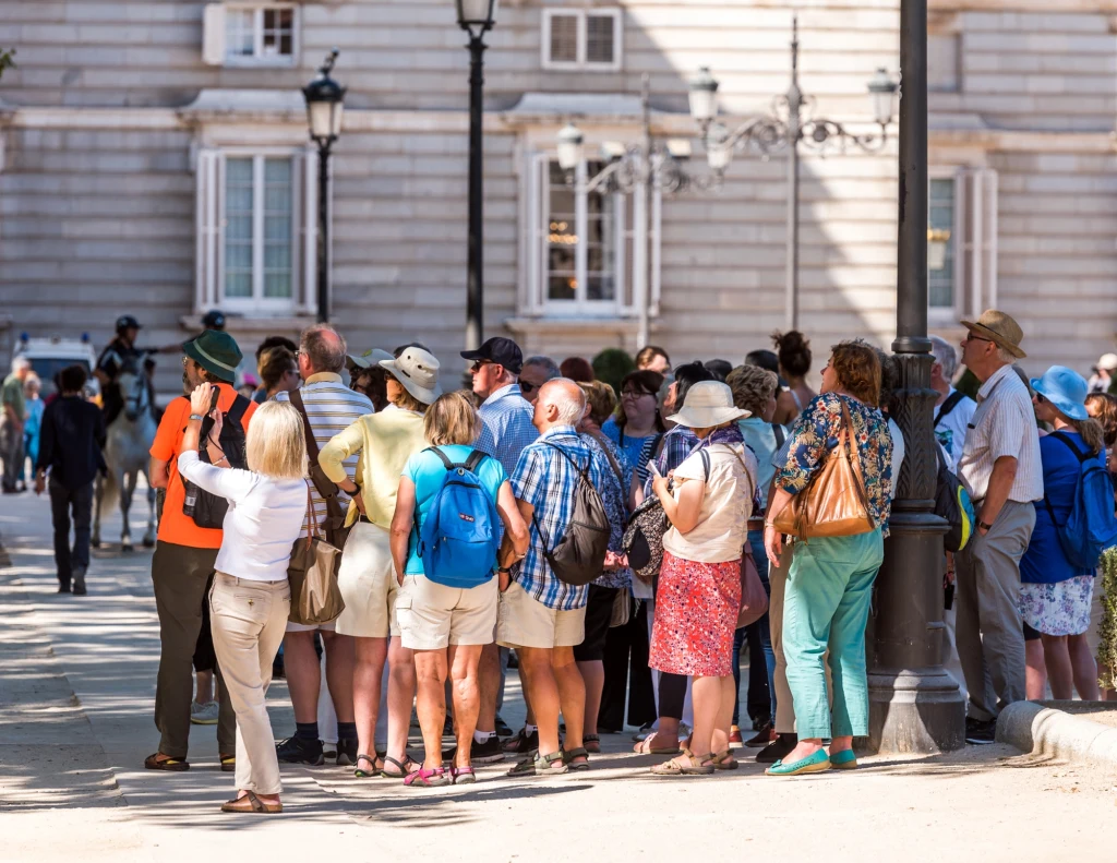 Zwiedzanie z przewodnikiem to szansa na lepsze poznanie historii i kultury danego miejsca Grupa turystów stoi na ulicy przed dużym, reprezentacyjnym budynkiem, większość osób ma na sobie letnie ubrania i kapelusze, część trzyma plecaki, jedna osoba robi zdjęcie grupie.