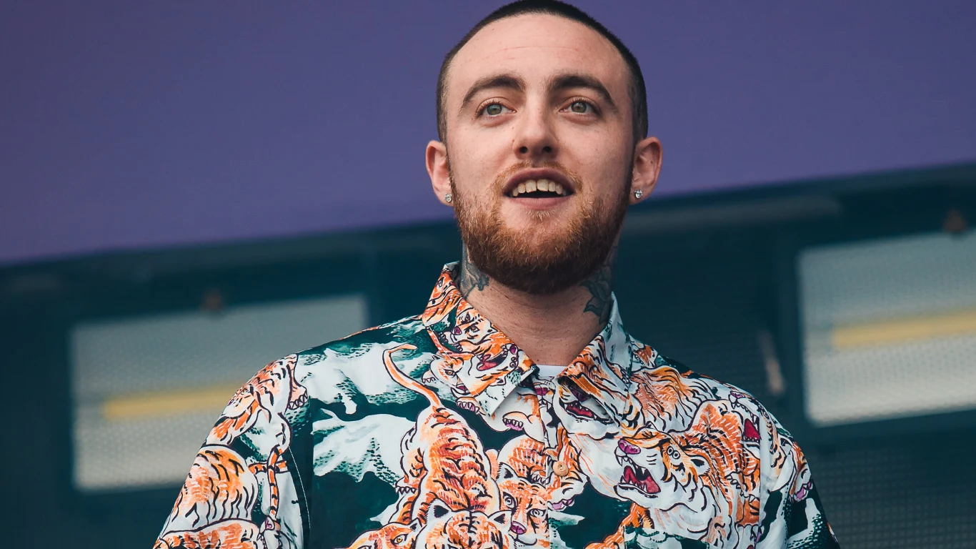 Mac Miller Młody mężczyzna z krótkimi włosami i brodą uśmiecha się lekko, ubrany w kolorową koszulę z nadrukiem tygrysów, stoi na tle fioletowej ściany i jasnych świateł.