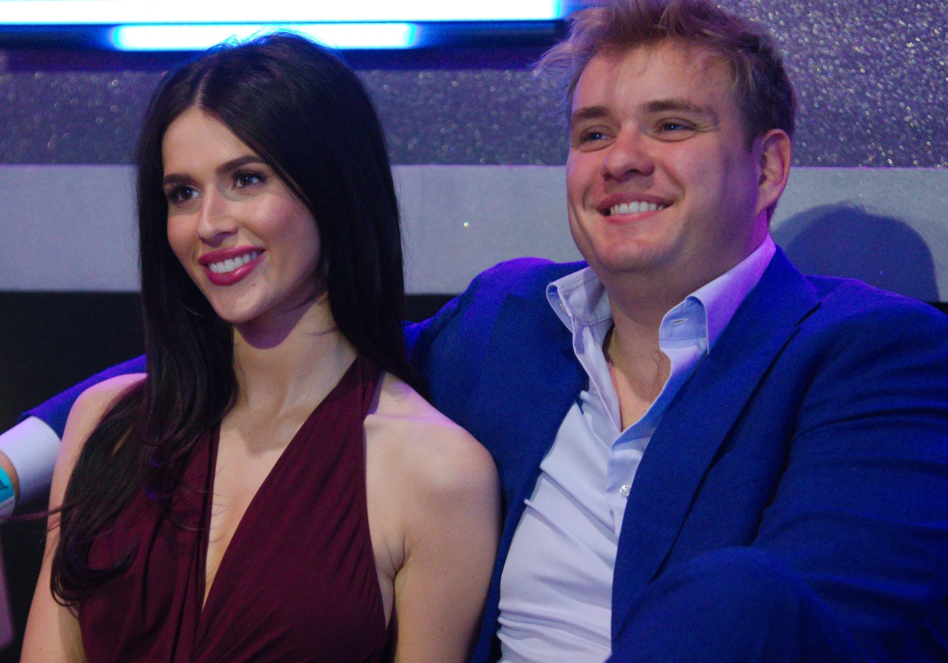 Roksana Węgiel i Kevin Mglej Para uśmiechniętych młodych ludzi siedząca razem, kobieta w bordowej sukni, mężczyzna w niebieskiej marynarce, oboje wyglądają na zadowolonych, tło w odcieniach niebieskiego.