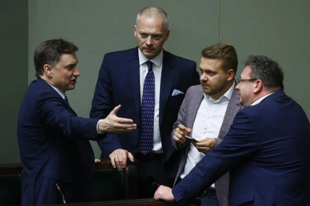 Czterech mężczyzn w garniturach uczestniczy w ożywionej dyskusji w jasnym pomieszczeniu, prawdopodobnie na sali sejmowej; jeden z nich gestykuluje ręką, pozostali słuchają uważnie.