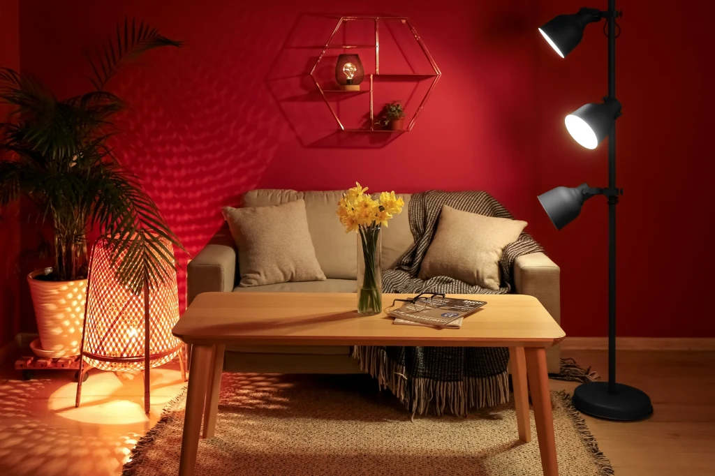 Odważny kolor może diameteralnie odmienić pokój Przytulny salon z czerwonymi ścianami, szarą sofą, drewnianym stolikiem i bukietem żółtych kwiatów w wazonie. Oświetlenie zapewnia stojąca lampa oraz ciepłe światło z lampy po lewej stronie, na ścianie wisząca ozdoba geometryczna.