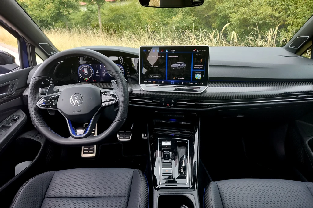 Poza kilkoma detalami wnętrze Golfa R jest takie samo, jak w zwykłych wersjach Nowoczesne wnętrze samochodu marki Volkswagen z cyfrowym zegarami, dużym dotykowym ekranem centralnym, ergonomicznie rozmieszczonymi przyciskami i eleganckimi siedzeniami, deska rozdzielcza o minimalistycznym wzornictwie, widok na otoczenie samochodu p...