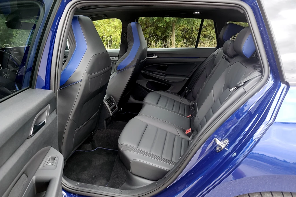 Z tyłu miejsca nie powinno brakować nikomu Wnętrze samochodu z widocznymi tylnymi siedzeniami pokrytymi czarną tapicerką, otwartymi drzwiami i detalami wykończenia o sportowym charakterze, częściowo widoczne przednie fotele oraz przestronne tylne miejsce na nogi.