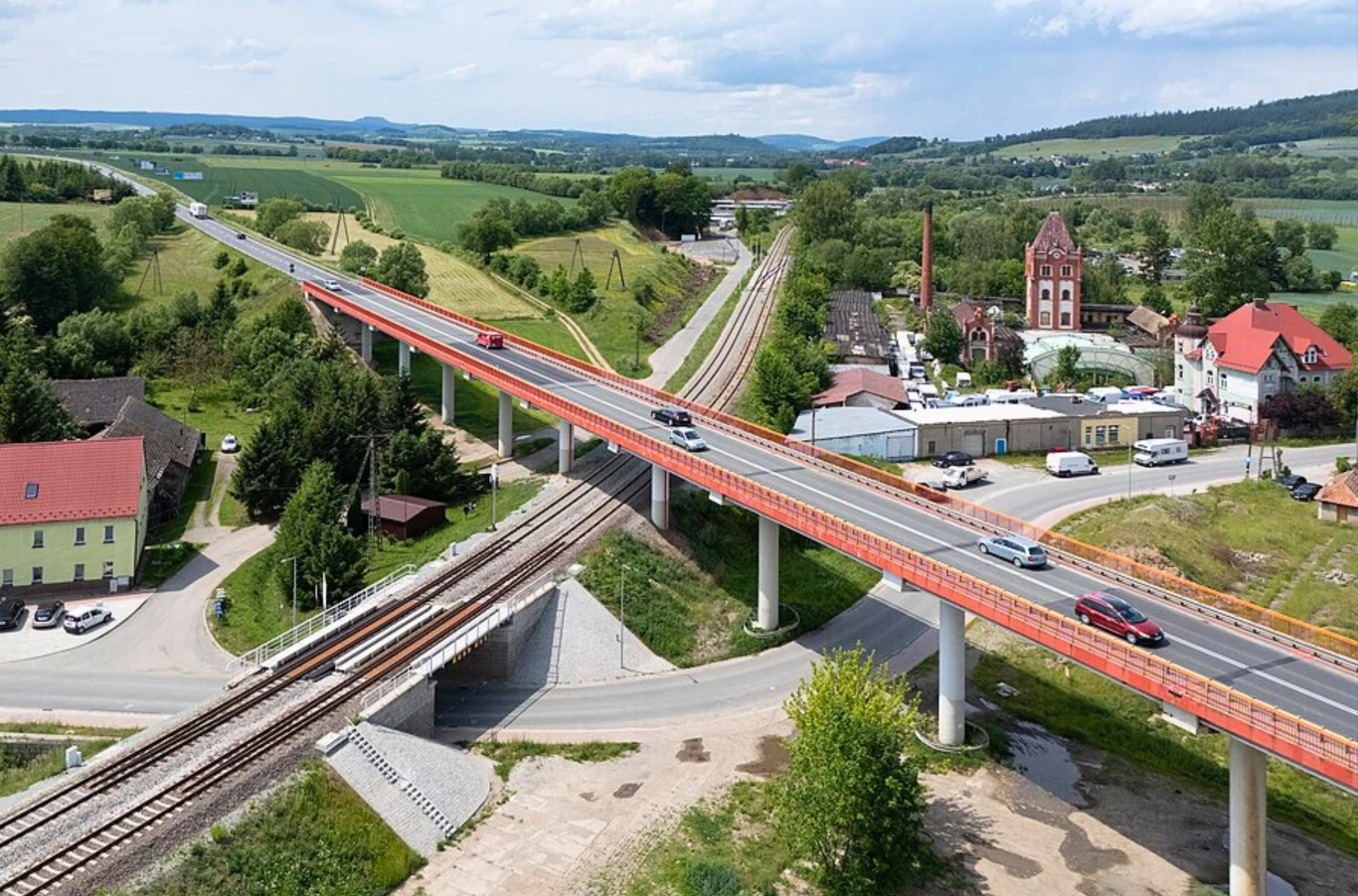 Od Czech po Podlasie. Oto najdłuższa droga krajowa w Polsce. Nowoczesny wiadukt drogowy przebiegający nad torami kolejowymi oraz skrzyżowaniem, otoczony zielonymi polami, zabudowaniami mieszkalnymi i przemysłowymi, z widocznymi samochodami poruszającymi się po moście.