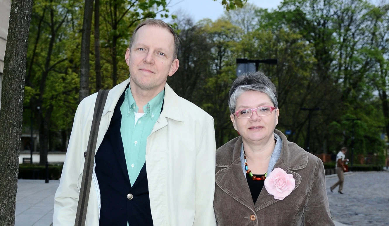 Dorota Zawadzka z mężem Dwoje dorosłych stoi razem w parku, otoczeni drzewami. Mężczyzna w jasnym płaszczu i kobieta w okularach, ubrana w brązowy płaszcz z różowym kwiatem, patrzą na obiektyw. W tle spacerują pojedyncze osoby.