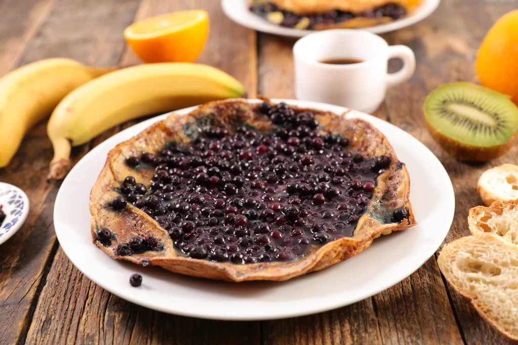 Owsianka zapieczona z borówkami podana na białym talerzu, otoczona świeżymi owocami takimi jak banan, kiwi i pomarańcza, filiżanka kawy oraz kawałki chleba na drewnianym stole.