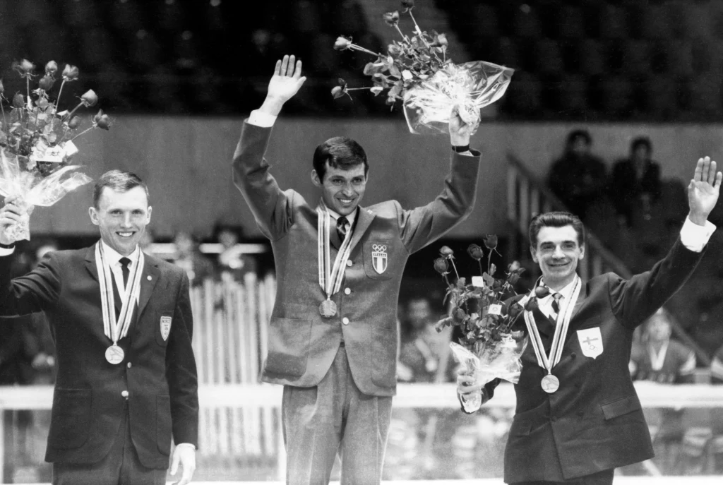 Trzech medalistów na podium, wszyscy trzymają bukiety kwiatów i mają medale na szyjach, środkowy zawodnik stoi na najwyższym stopniu, wszyscy uśmiechnięci i machają do publiczności.