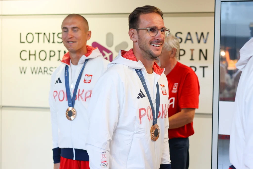 Dwóch uśmiechniętych sportowców ubranych w białe bluzy z napisem 'Polska', mających na szyjach medale, stojących w hali lotniska Chopina w Warszawie; w tle osoba w czerwonej koszulce.