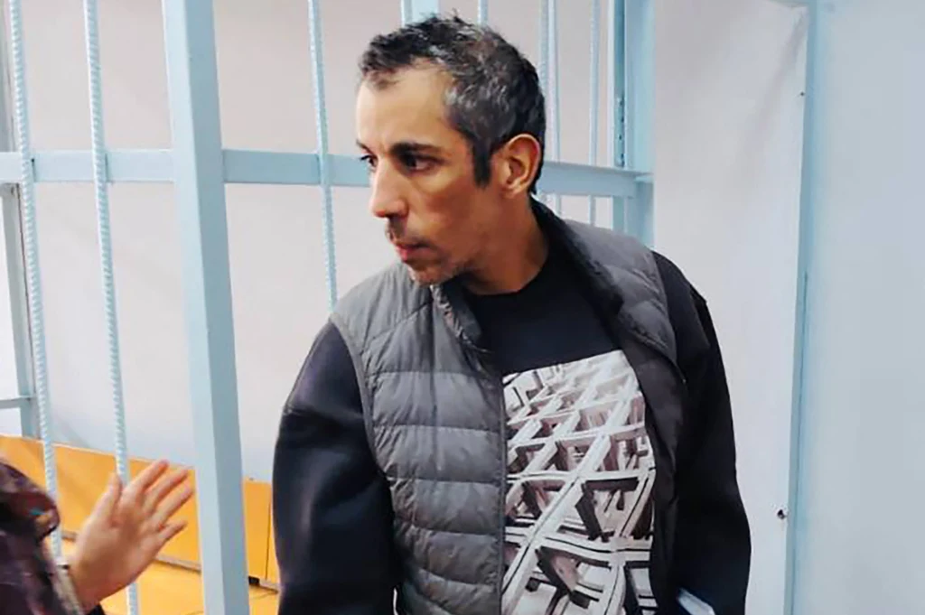 Mężczyzna o ciemnych włosach i zarostem stoi w pomieszczeniu z metalowymi kratami, ubrany w ciemną ocieplaną kamizelkę i czarną koszulkę z nadrukiem. W tle widoczne są metalowe słupki i ławka.