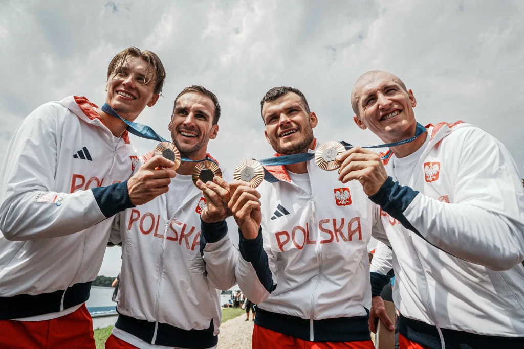 Czterech uśmiechniętych sportowców w biało-czerwonych dresach z napisem Polska prezentuje medale zdobyte podczas zawodów sportowych. Grupa stoi razem na tle nieba, trzymając medale na niebieskich wstążkach.