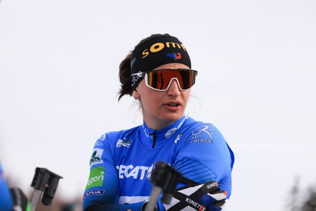 Kobieta ubrana w sportową niebieską odzież, opaskę z napisem i okulary przeciwsłoneczne trzyma kijki do narciarstwa biegowego, stoi na tle jasnego nieba.