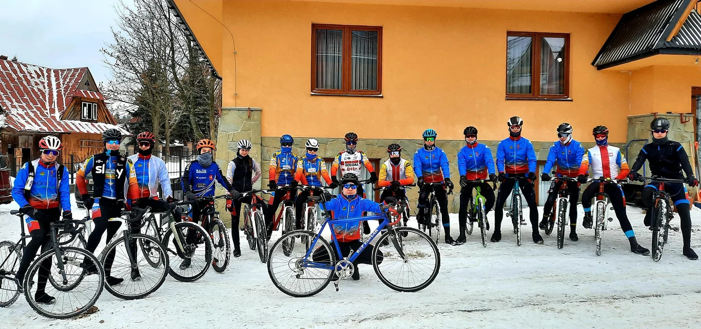 Grupa rowerzystów ubranych w niebiesko-czerwone stroje stoi z rowerami na pokrytym śniegiem podłożu, na tle pomarańczowego budynku z dwoma oknami i widocznym dachem. W centralnej części jedna osoba siedzi na rowerze, reszta stoi lub opiera się o rowery.